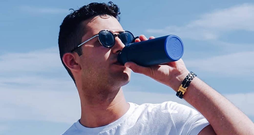 A male brand ambassador for Ocean Bottle drinks out his dark blue bottle on a summers day. He has short, dark curly hair, wears a white t-shirt, dark sunglasses with a watch on his wrist.