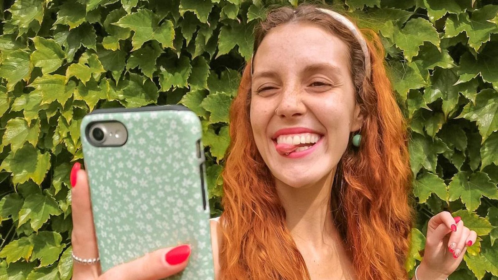 A red haired Burga ambassador holds out her phone while taking a selfie. She is winking and her tongue is stuck out. Her mint green phone case is dotted with flowers but not in focus.
