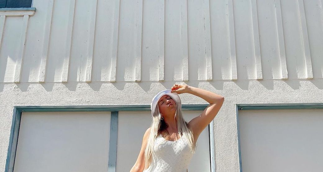 A DippinDaisys ambassador wears a white bucket hat and a white full swimpiece. She stands in front of a textured white wall while holding her hat and pointing her face up toward the sun.
